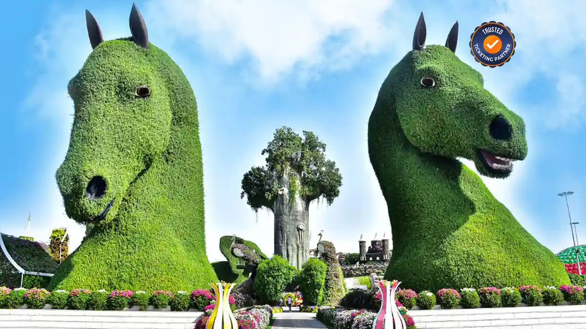 Two large, green topiary sculptures shaped like horse heads dominate a vibrant garden scene. Bright flowers surround them under a clear blue sky.