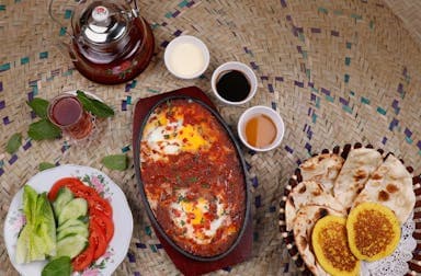 A vibrant Middle Eastern meal on a woven mat. Eggs in tomato sauce, assorted breads, sliced vegetables, sauces, and tea 