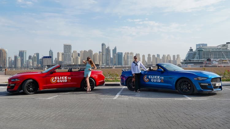 Two convertible cars, one red and one blue, parked side by side with the backdrop of a city skyline. 