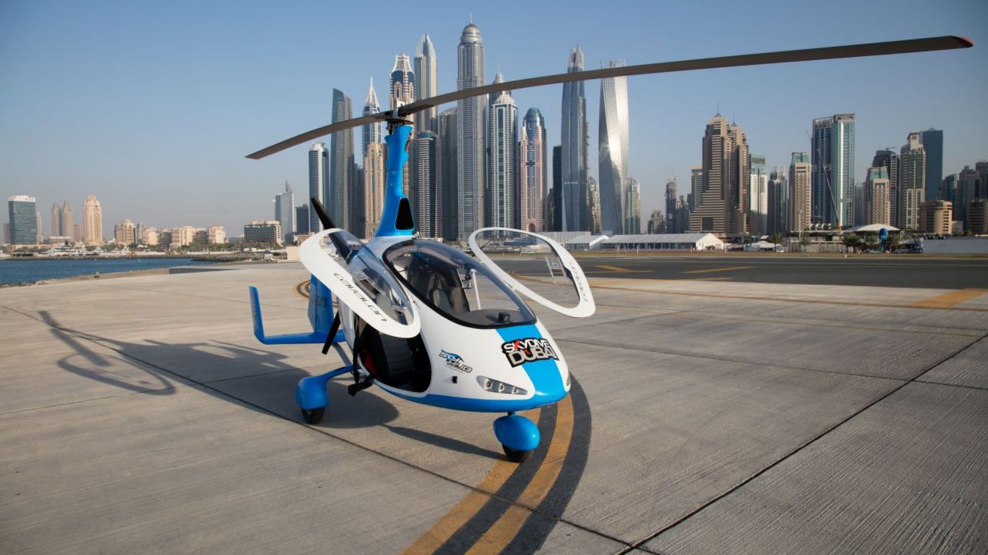 A gyrocopter on the tarmac with a city skyline in the background, labeled as the Gyrocopter Flight Helipad.