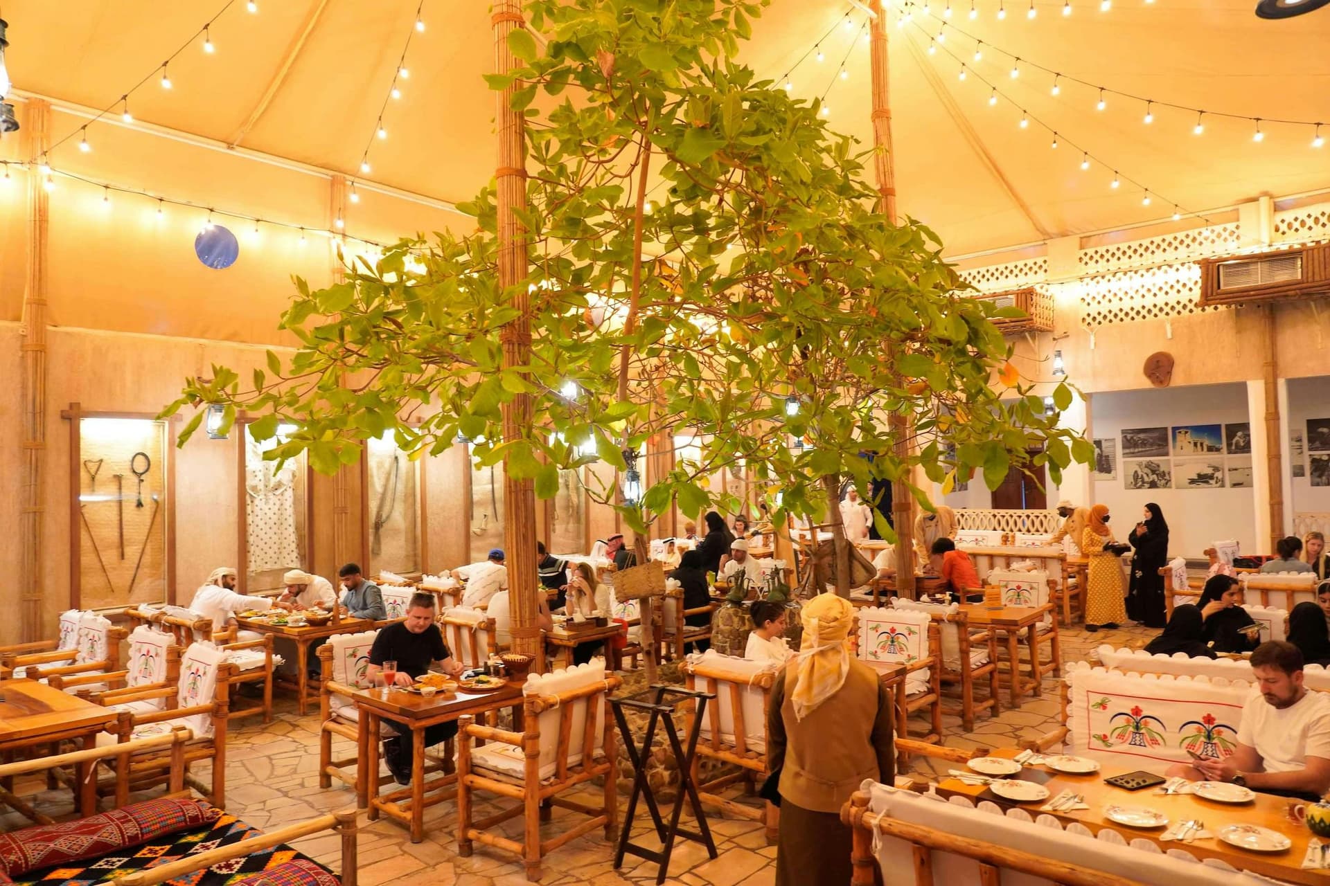 A warmly lit, cozy restaurant with a tree at its center, surrounded by wooden tables and chairs. 