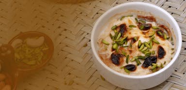 A white bowl of creamy dessert topped with toasted nuts and sliced pistachios on a woven mat. Warm and inviting tones suggest a cozy atmosphere.