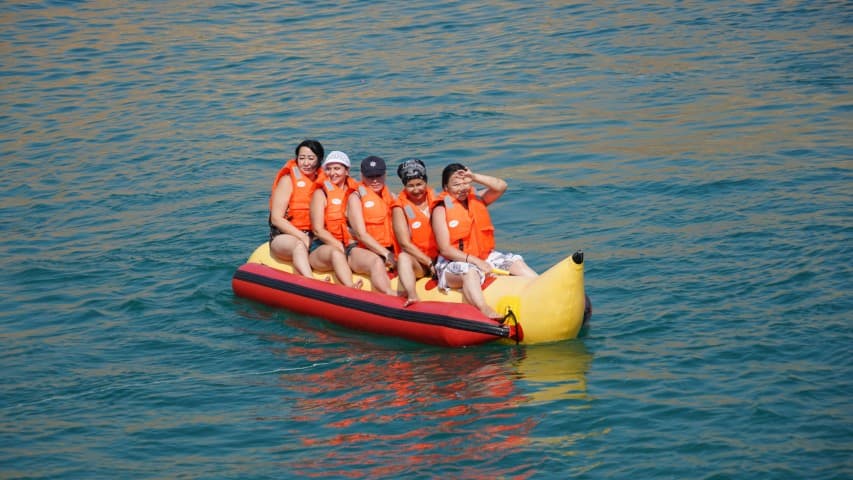 Five people wearing orange life vests joyfully ride a yellow inflatable banana boat on clear blue water