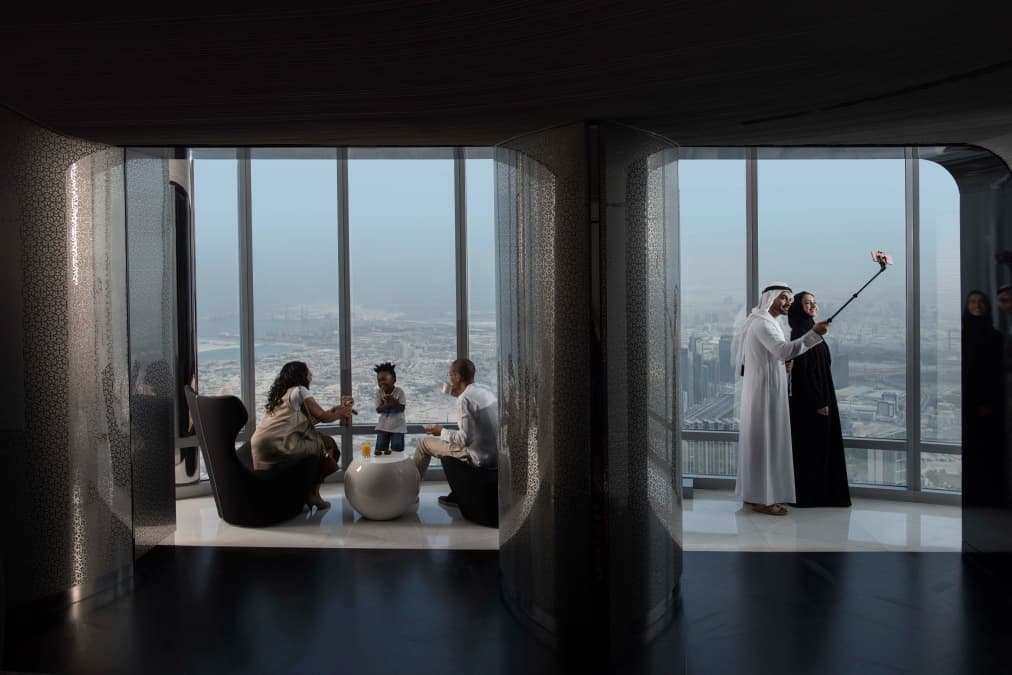 Two groups enjoy a scenic city view from the Burj Khalifa. On the left, a family sits chatting. On the right, a couple in traditional attire takes a selfie.