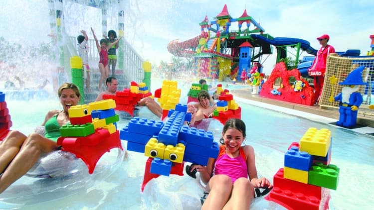 Children and adults joyfully float on colorful inflatable Lego pieces in a water park