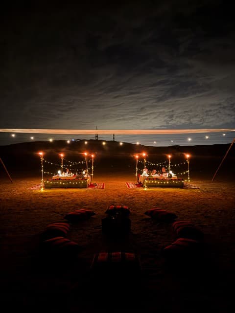 A couple of tents illuminated under the night sky in the desert, set up for a camp dinner.