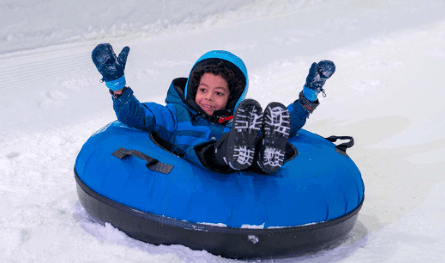 Snow Abu Dhabi: A child in a blue snowsuit and gloves joyfully rides a blue snow tube down a snowy hill, smiling with raised arms