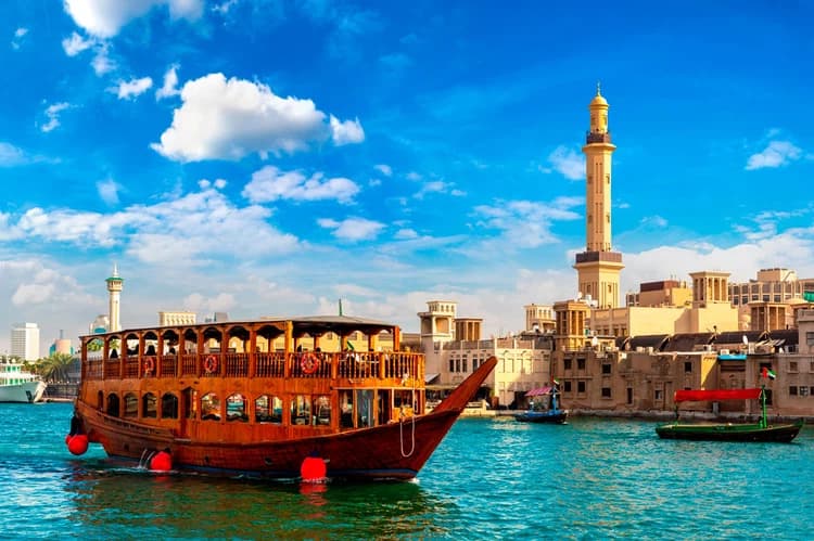 A traditional wooden dhow boat floats on Dubai Creek under a vibrant blue sky. 