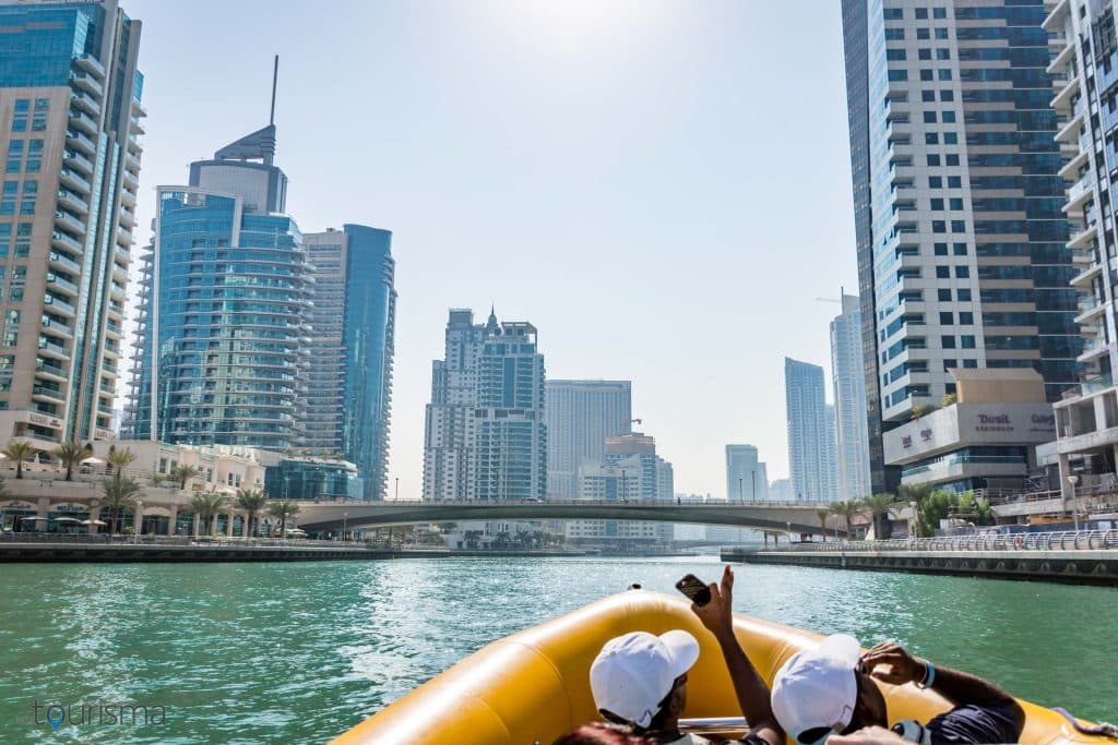 A boat glides along a waterfront, framed by tall modern skyscrapers in a sunny cityscape.