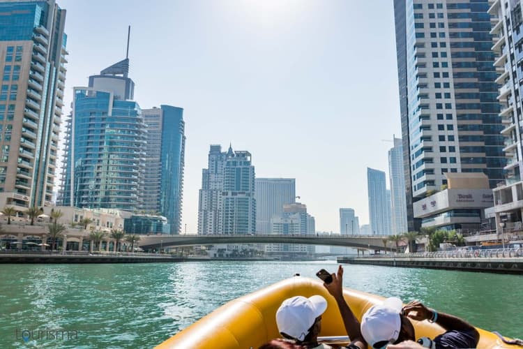 A boat glides along a waterfront, framed by tall modern skyscrapers in a sunny cityscape.