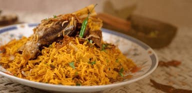 A plate of biryani topped with lamb shank and garnished with green chilies and herbs, set on a woven mat, creating a warm, appetizing ambiance.