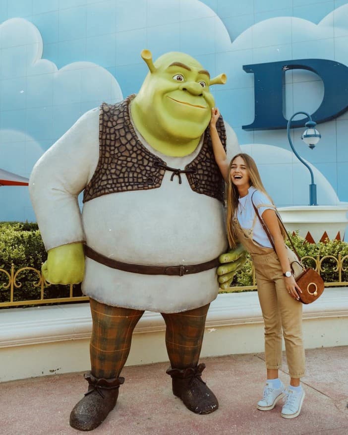 A woman playfully poses with a large Shrek statue, smiling and gently pinching its cheek