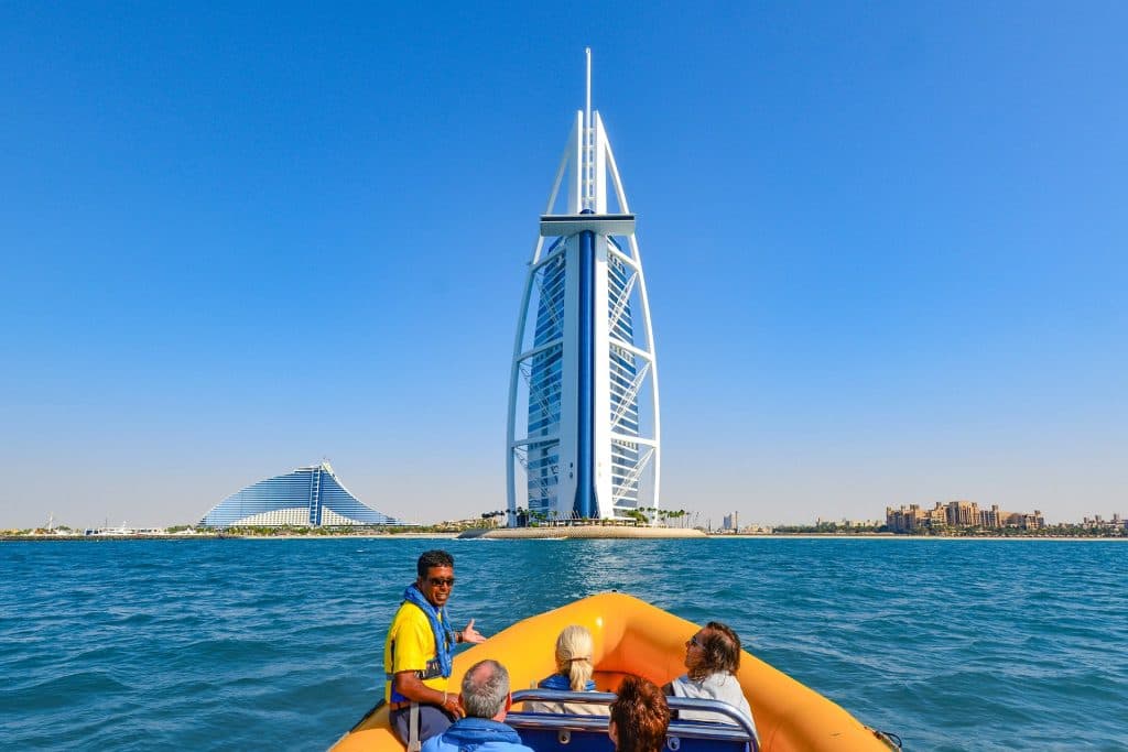 A group in an orange boat approaches a futuristic sail-shaped skyscraper on a sunny day,