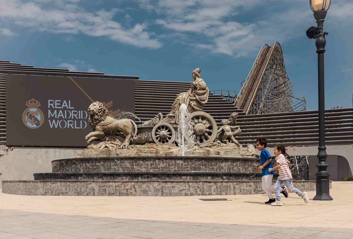 Two children run joyfully around A large fountain with a classical statue near a "Real Madrid World" sign.