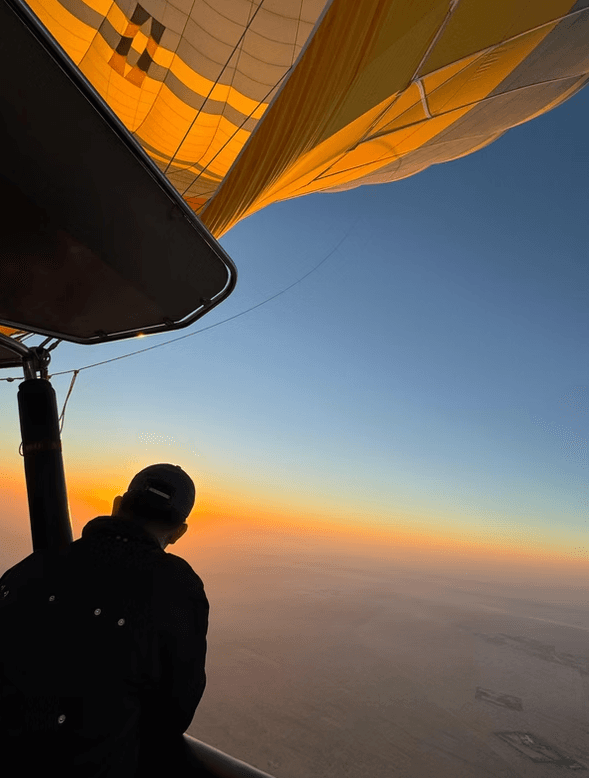 Silhouette of a person in a hot air balloon at sunrise, with an orange and blue horizon. 