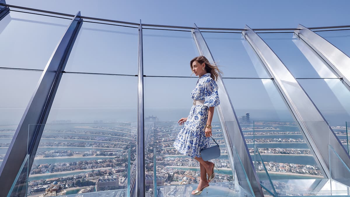 A woman looks out over the view at the palm while standing on a transparent glass floor high above.