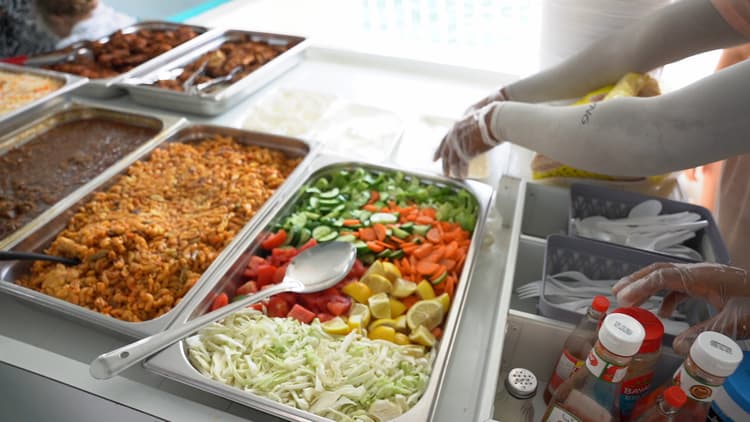 Buffet with trays of colorful salads, pasta, and sauces. A person with gloved hands arranges items