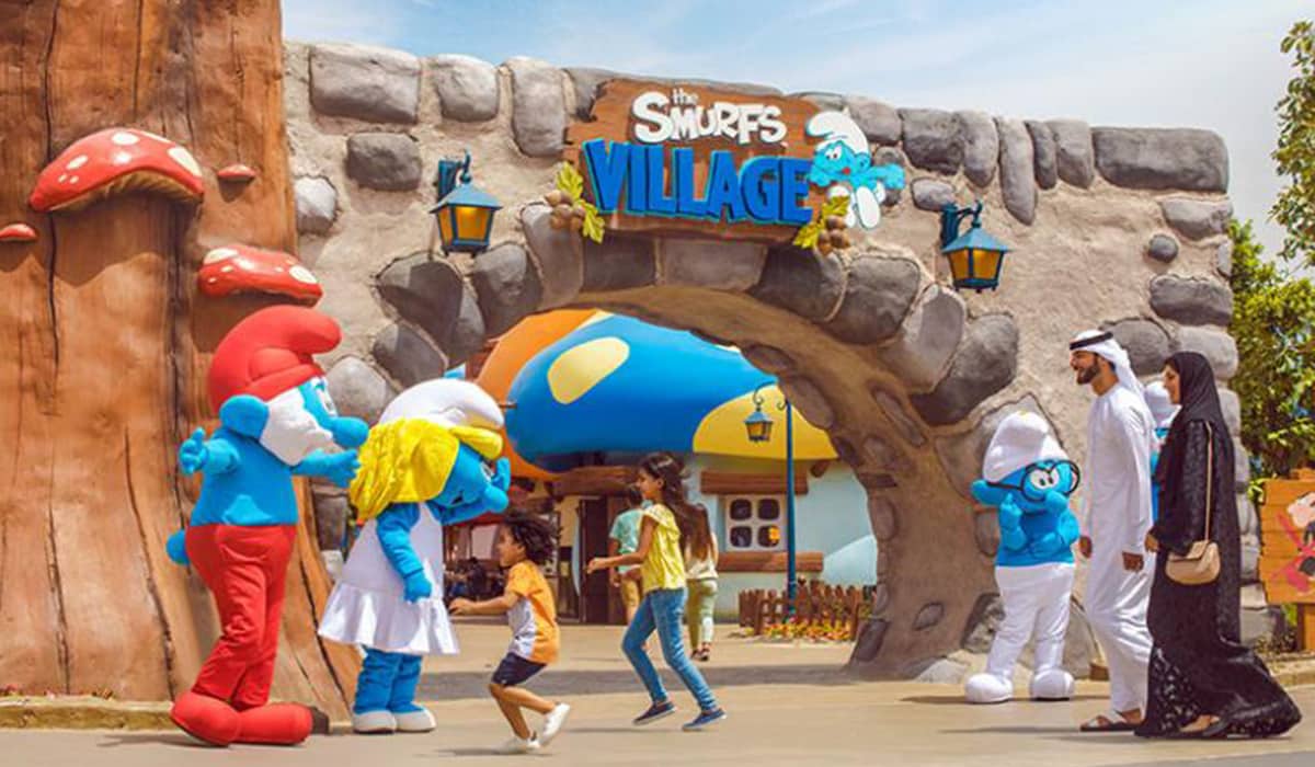 Children, excited and playful, walk with two adults, at a theme park enter Smurfs Village, greeted by Smurf mascots