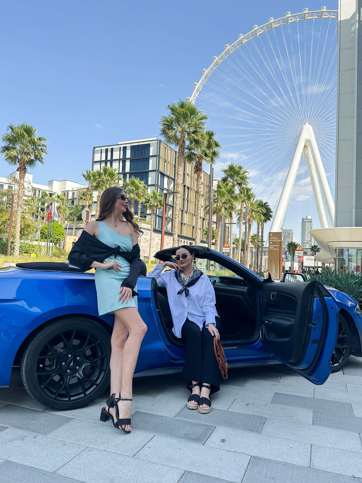 Two women in stylish outfits stand by a blue convertible car. 