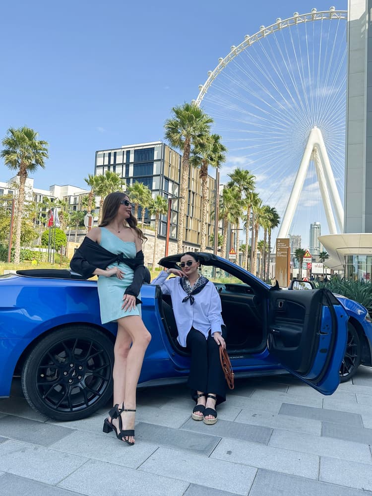 Two women in stylish outfits stand by a blue convertible car. 