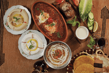 A Middle Eastern breakfast spread on a woven mat, featuring dishes like eggs in tomato sauce, hummus, breads, vegetables, and tea