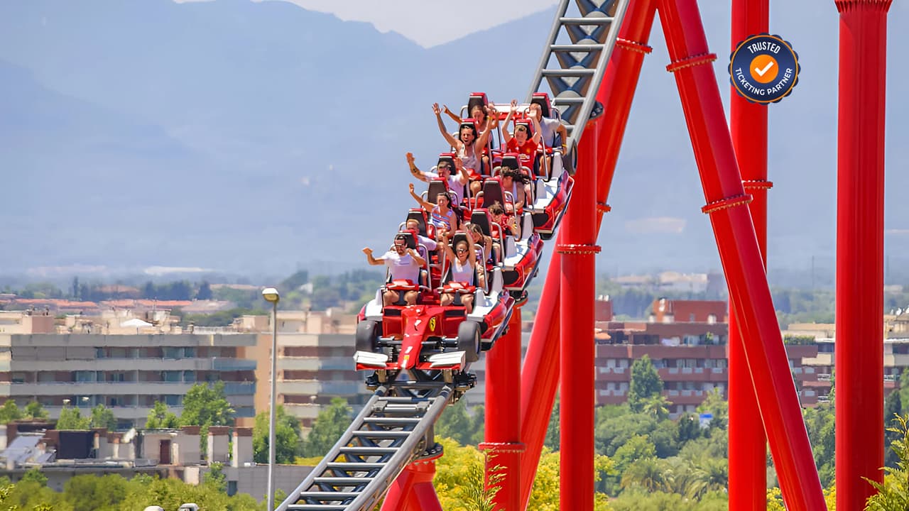 A group of people riding a red roller coaster at Ferrari World Abu Dhabi, descending a steep track with mountain and cityscape in the background.