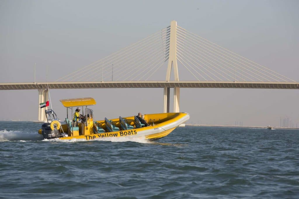 A yellow boat with people aboard floats in the water, set against the backdrop of Abu Dhabi City.