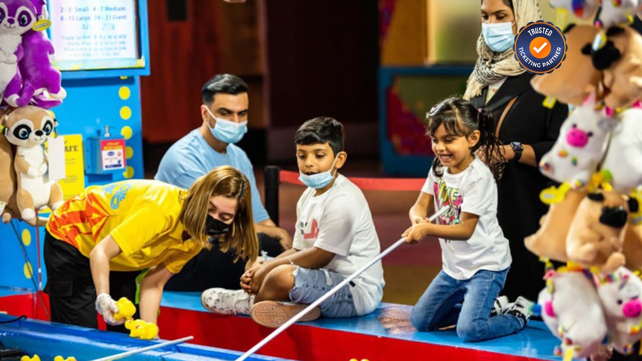 Children play a duck pond game at a fair, guided by an attendant. They look happy and engaged.