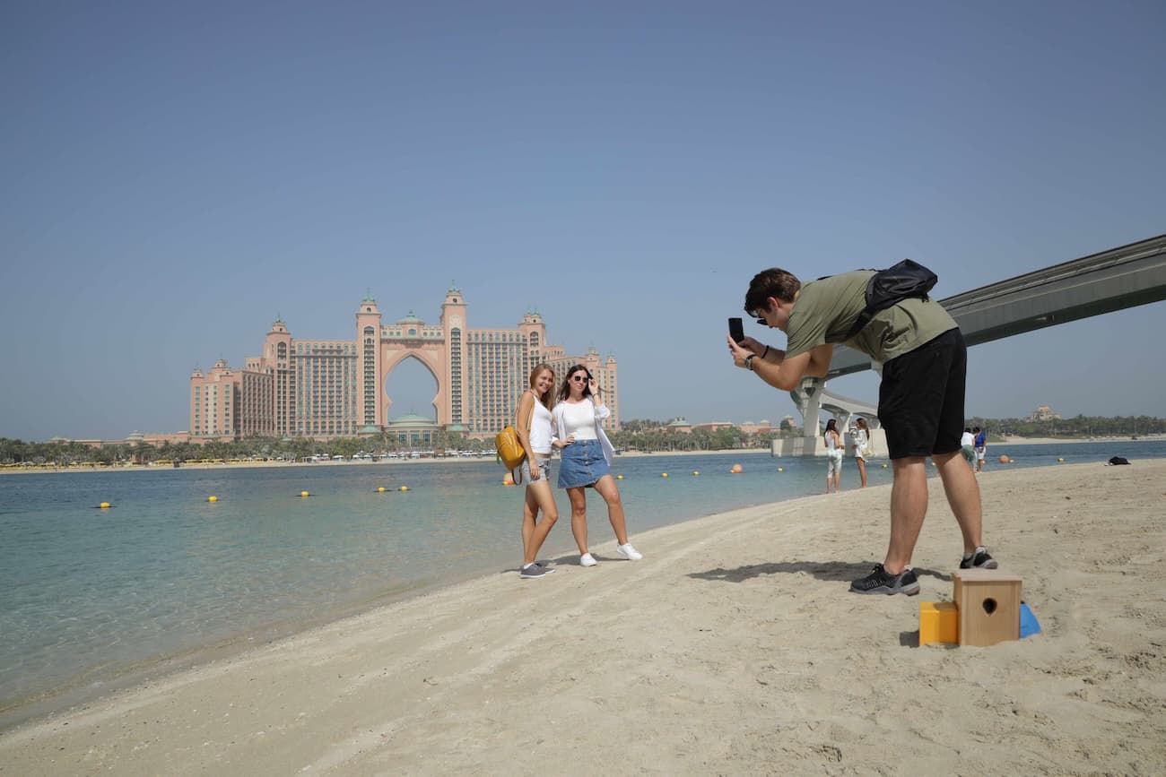 Two women pose on a sunny beach near a clear blue shoreline, with a grand hotel in the background.