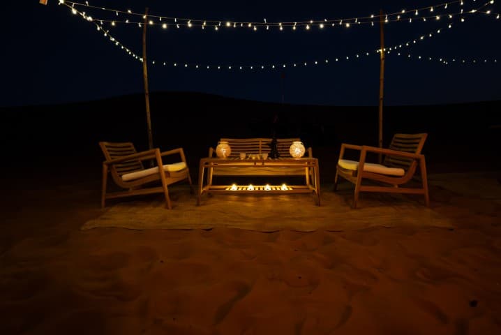 Outdoor desert setting with wooden chairs around a glowing firepit. String lights above create a cozy, intimate atmosphere.