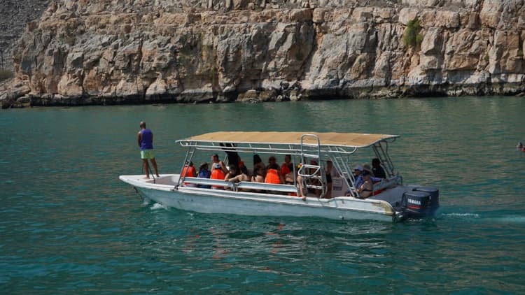 A boat with a beige canopy carries passengers in orange life vests on turquoise water.