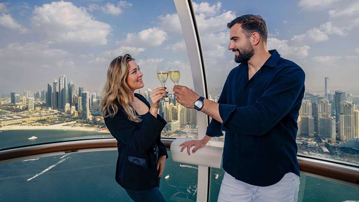 Couple celebrating with drinks inside an Ain Dubai cabin overlooking Dubai Marina skyline and the Arabian Gulf.
