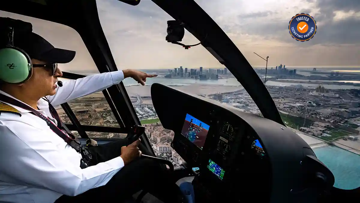 A helicopter pilot, wearing a white shirt and green headset, points toward a city skyline over a coastal landscape. The cockpit is in the foreground.