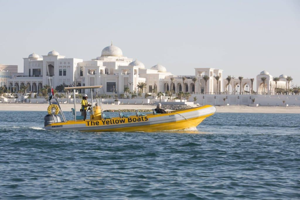 A yellow and white boat floats in the water near Qasr Al Watan, a large building in Abu Dhabi.
