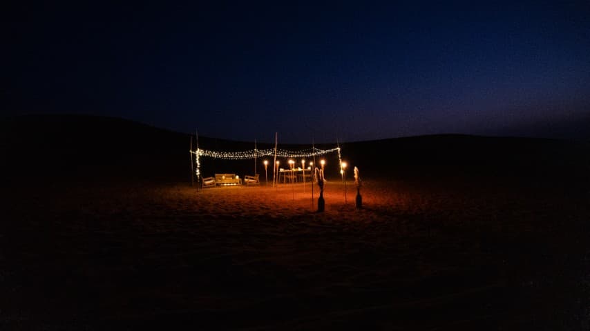 Nighttime desert scene with a lit-up canopy featuring string lights and torches. A lone bench sits under the canopy, casting a warm, inviting glow.