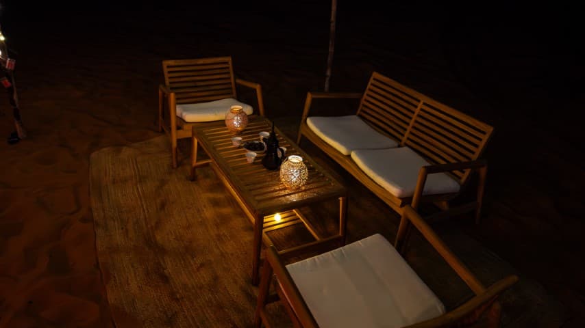 Outdoor wooden furniture set with white cushions arranged around a table holding decorative lanterns, set against a nighttime sandy backdrop, creating a serene and cozy atmosphere.