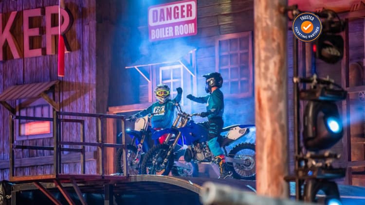Two motorcyclists in vibrant gear appear ready to race on a wooden platform, surrounded by theatrical smoke and stage lighting.