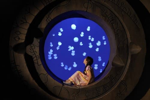 A girl gazes at floating bubbles from a round window, surrounded by the vibrant Aqua Trek Xtreme Jelly Fish exhibit.